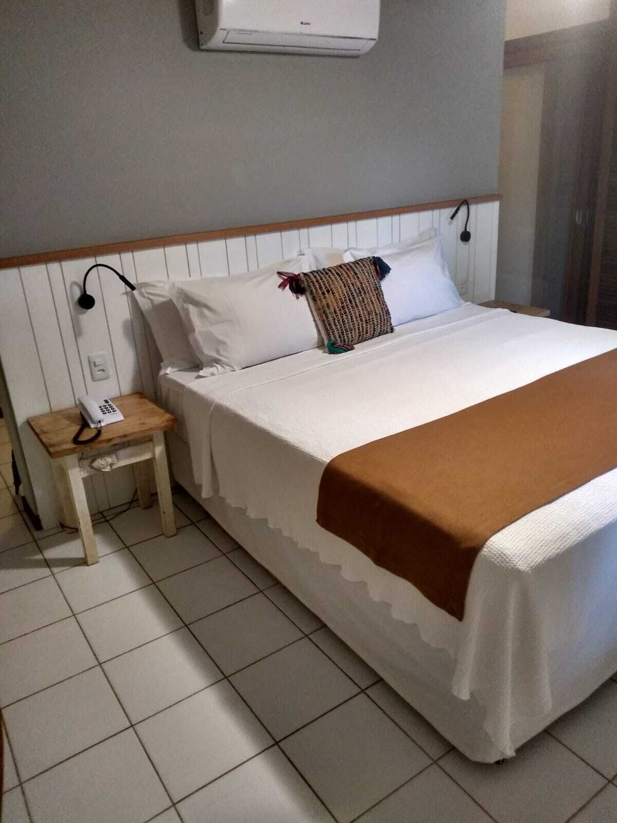 Un letto rifatto con lenzuola bianche e una coperta marrone, un comodino con un telefono, e un condizionatore d'aria a parete.