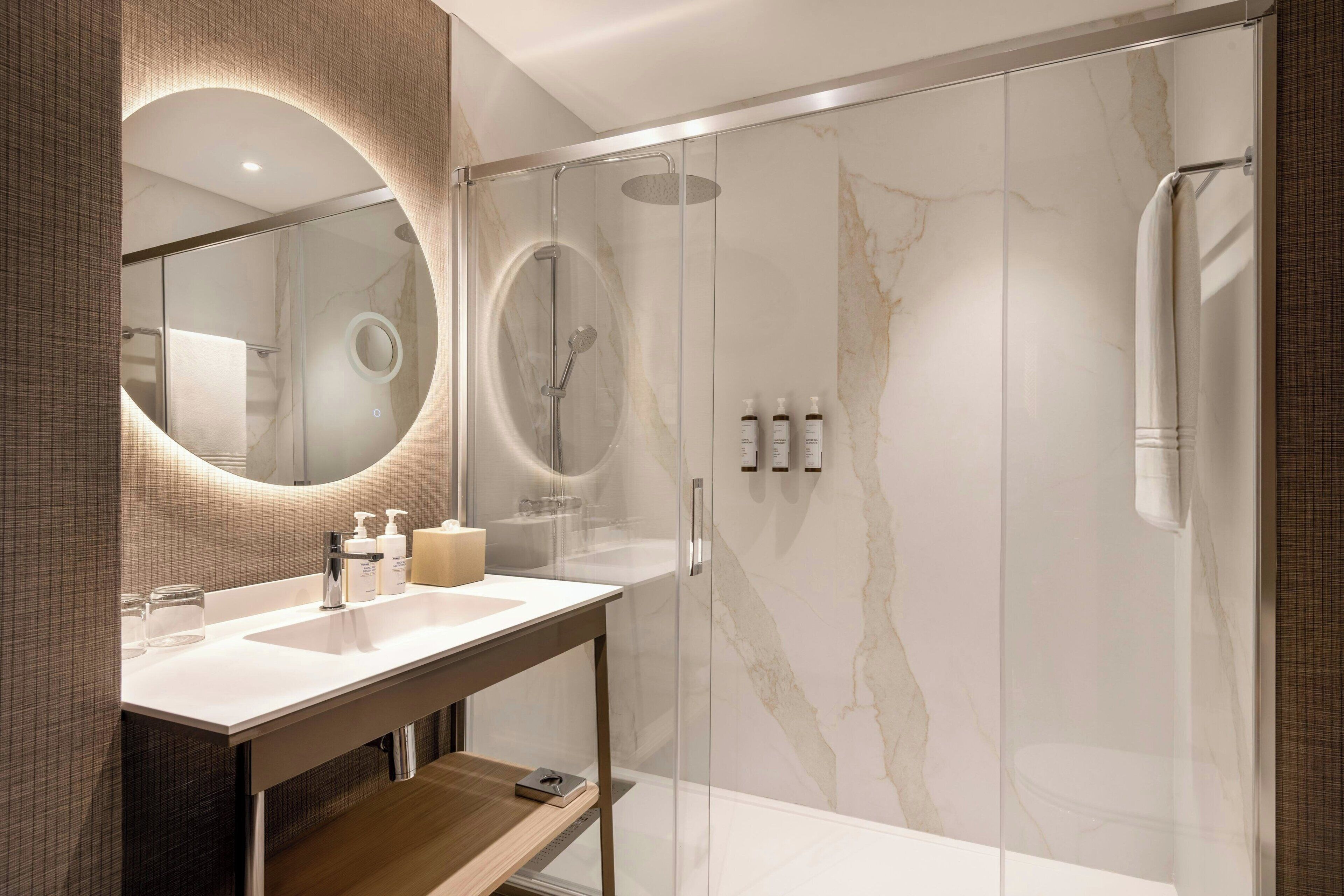 A modern bathroom with a glass-enclosed shower, a marble wall, and a vanity with a mirror and toiletries.