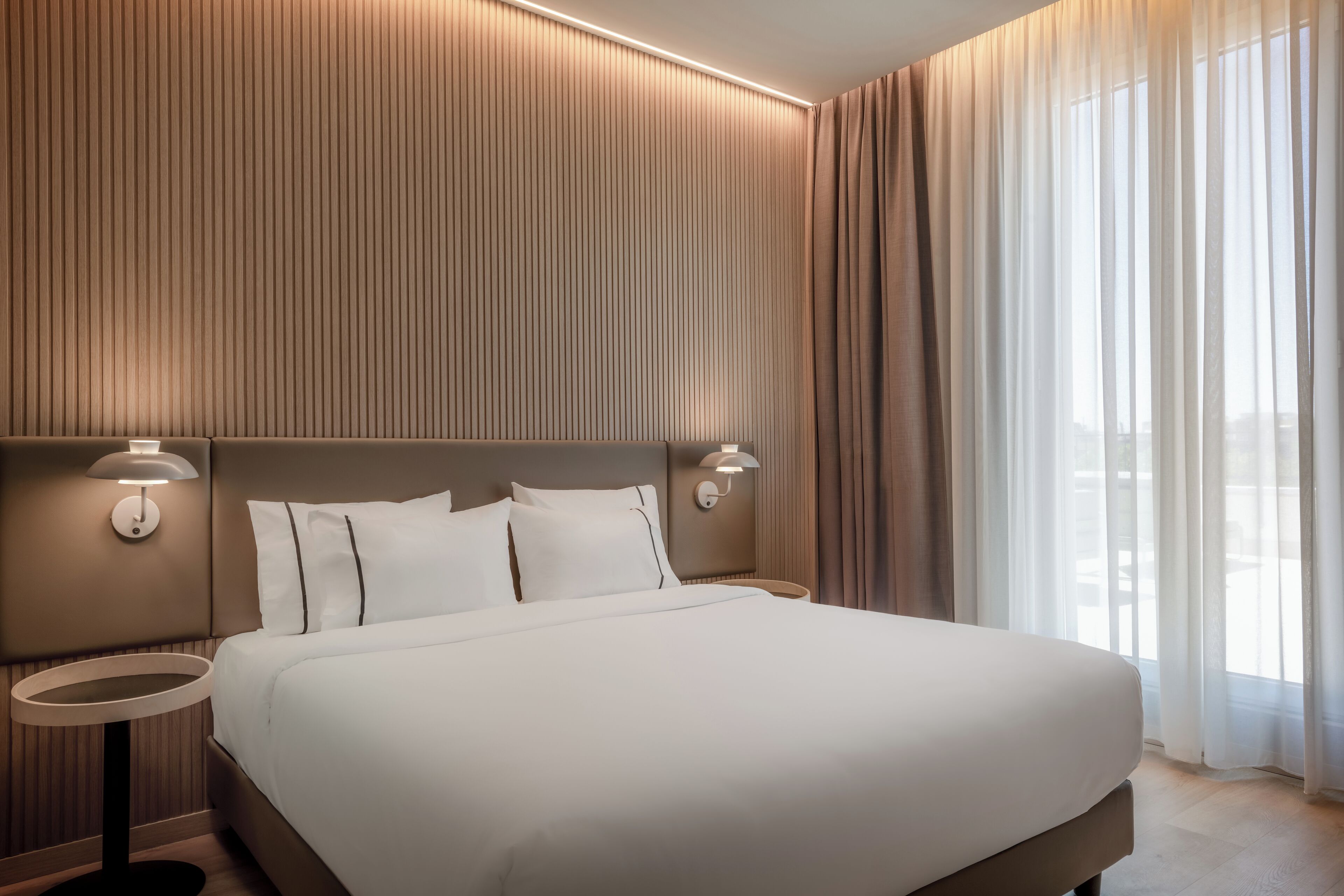 A modern hotel room with a large bed, two bedside tables, and a view of the outdoors through sheer curtains.