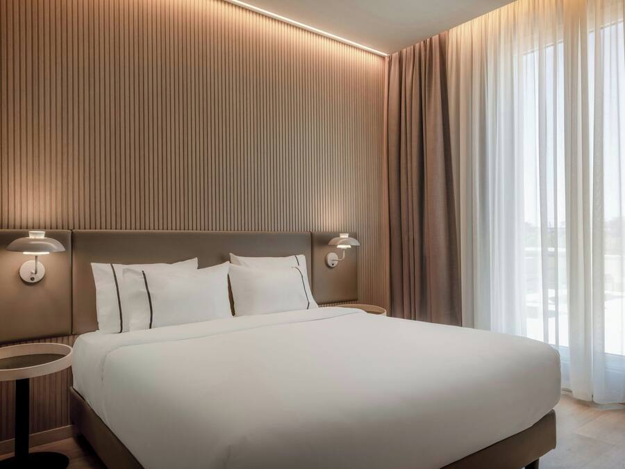 A modern hotel room with a large bed, two bedside tables with lamps, a wall with vertical lines, and a view through sheer curtains.