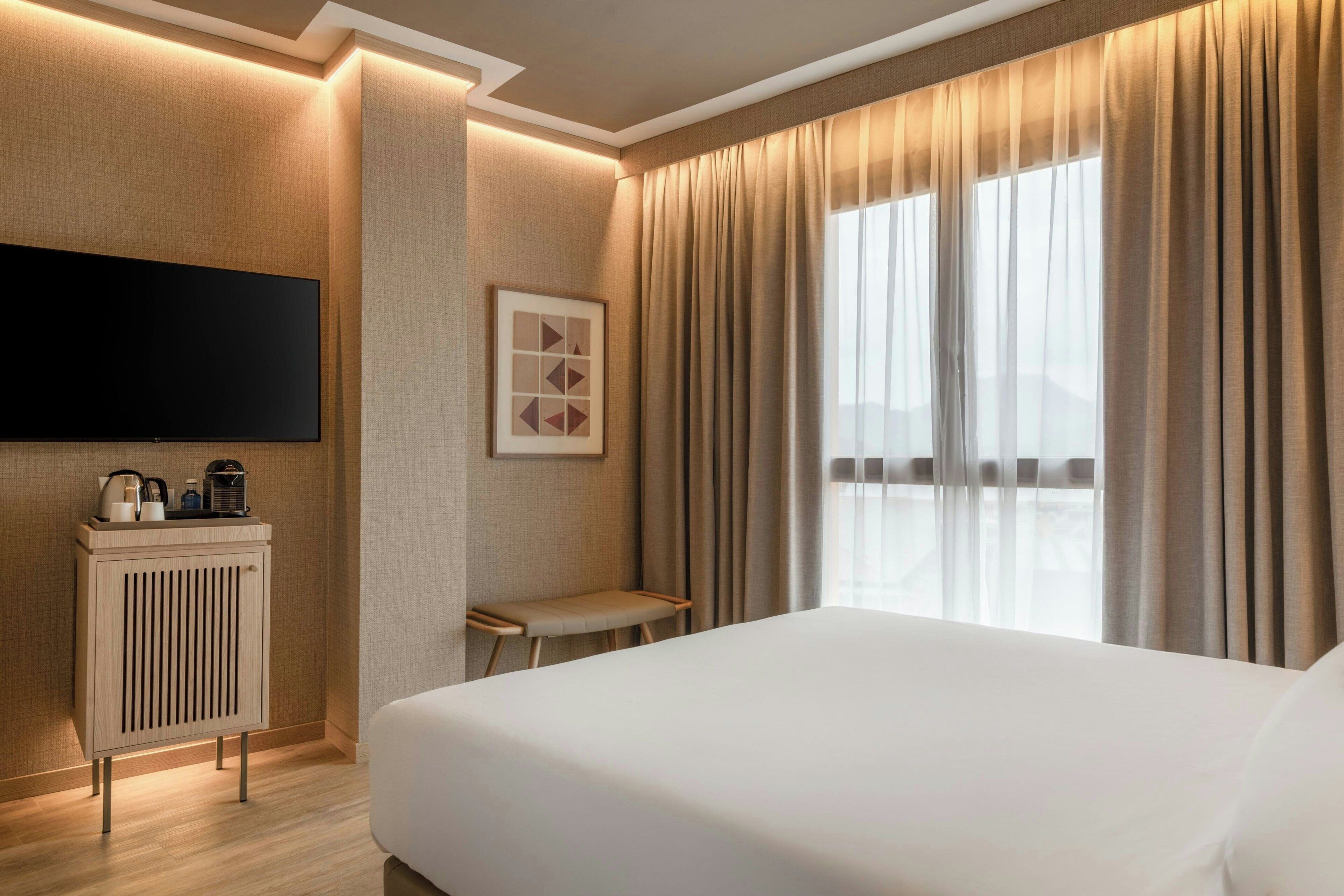 A hotel room with a large bed, a flat-screen TV mounted on the wall, a small table with a chair, and a window with curtains.