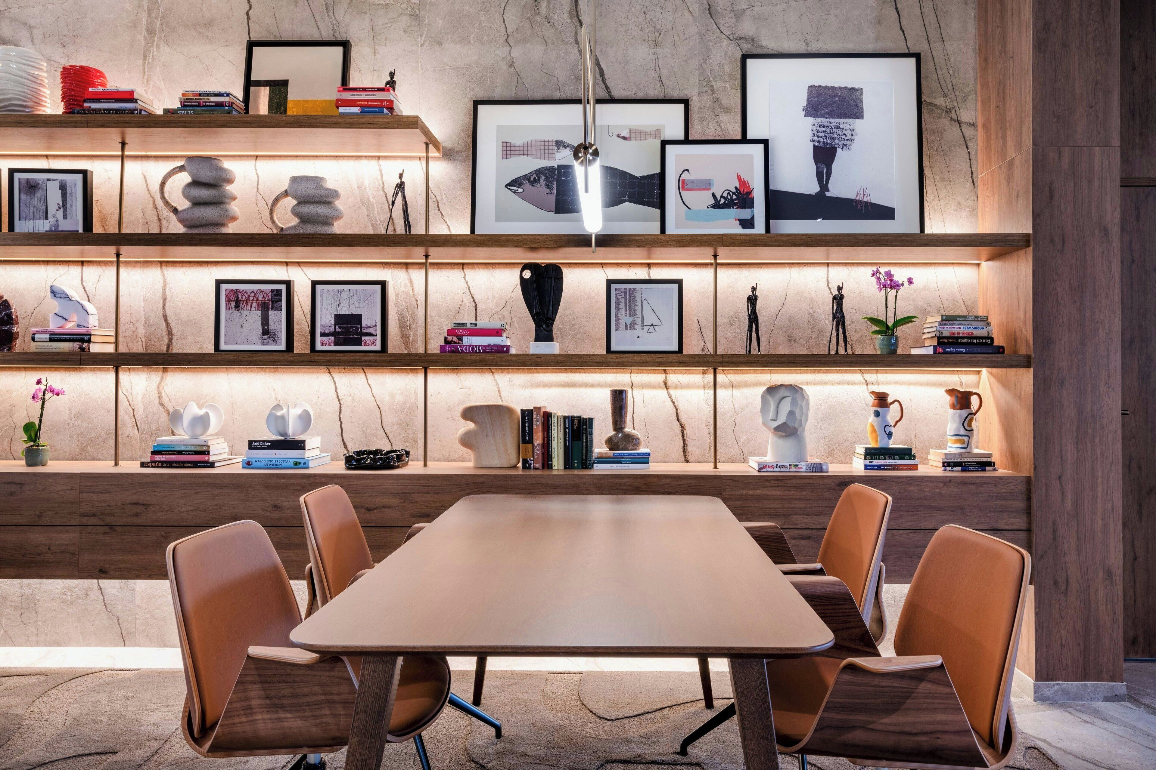 Un interior moderno con una mesa y sillas de madera, estanterías con libros y objetos decorativos, y una pared de mármol.
