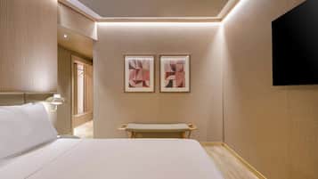 Une chambre d’hôtel moderne avec un grand lit, un banc, un téléviseur fixé au mur et deux œuvres d’art encadrées.
