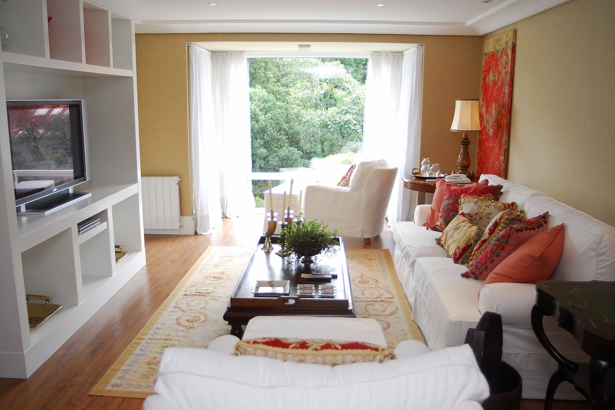 A living room with a white sofa, a coffee table, a TV, and a view of greenery outside.