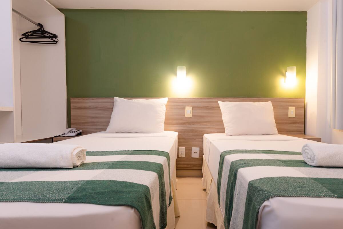 Habitación de hotel con dos camas individuales, cada una con una colcha verde y blanca a rayas, almohadas blancas y cabeceros de madera. Hay luces fijadas en la pared sobre cada cama y un teléfono en la mesita de noche.