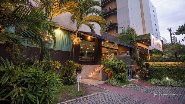 A modern hotel with a glass facade, outdoor seating area, and lush greenery. - Rede Andrade Vela Branca (Recife)