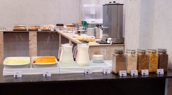 Un bufé con diversos alimentos y bebidas, como melones, papaya, leche, yogur, chocolate en polvo, avena, granola y galletas.