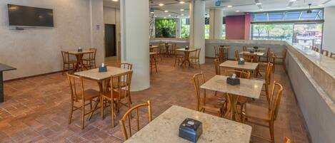 Un amplio comedor interior con múltiples mesas y sillas, un televisor de pantalla plana montado en la pared y grandes ventanales que proporcionan luz natural.