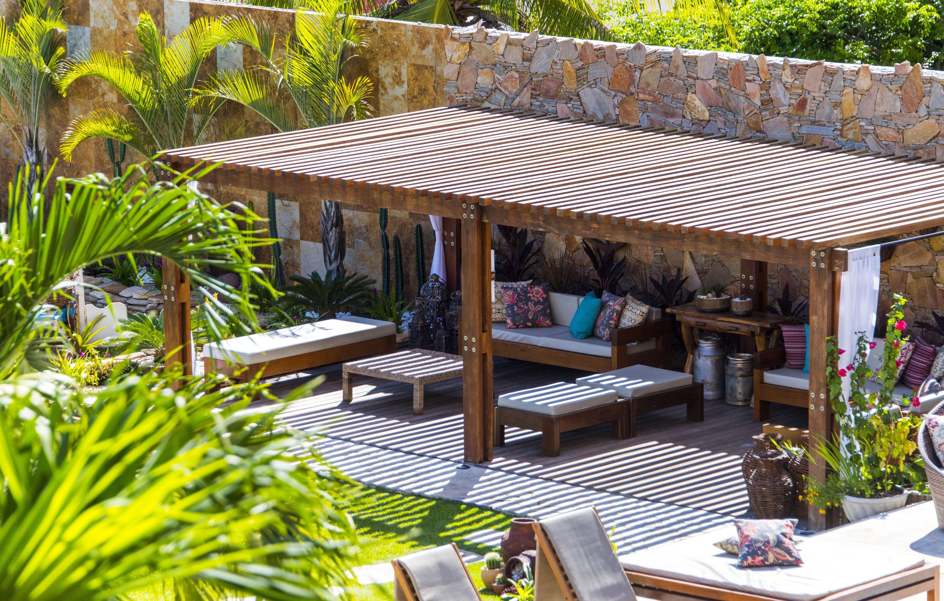 A covered outdoor seating area with wooden benches and cushions, surrounded by potted plants and a stone wall.