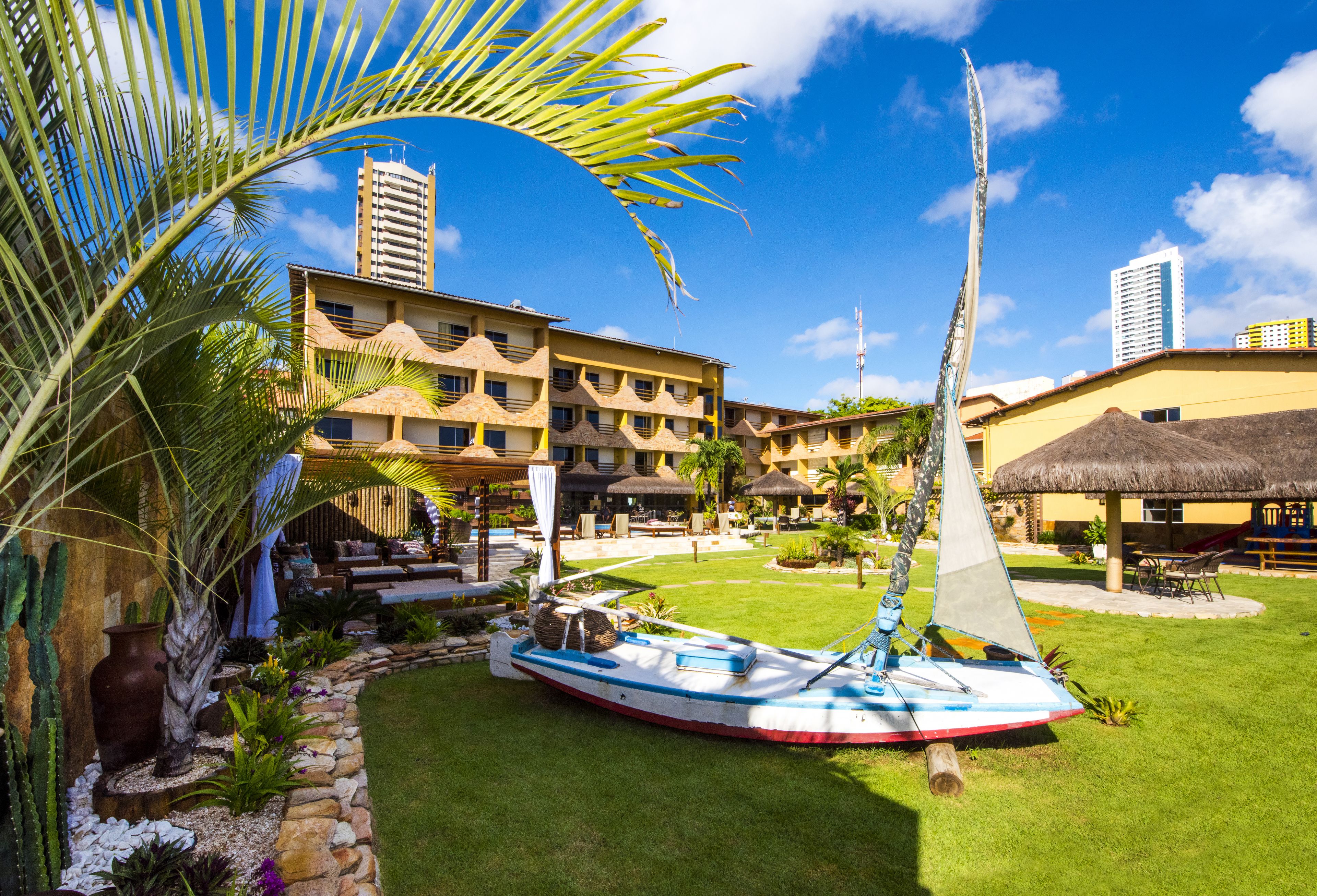 A resort with a swimming pool, a sailboat, and a thatched-roof building.
