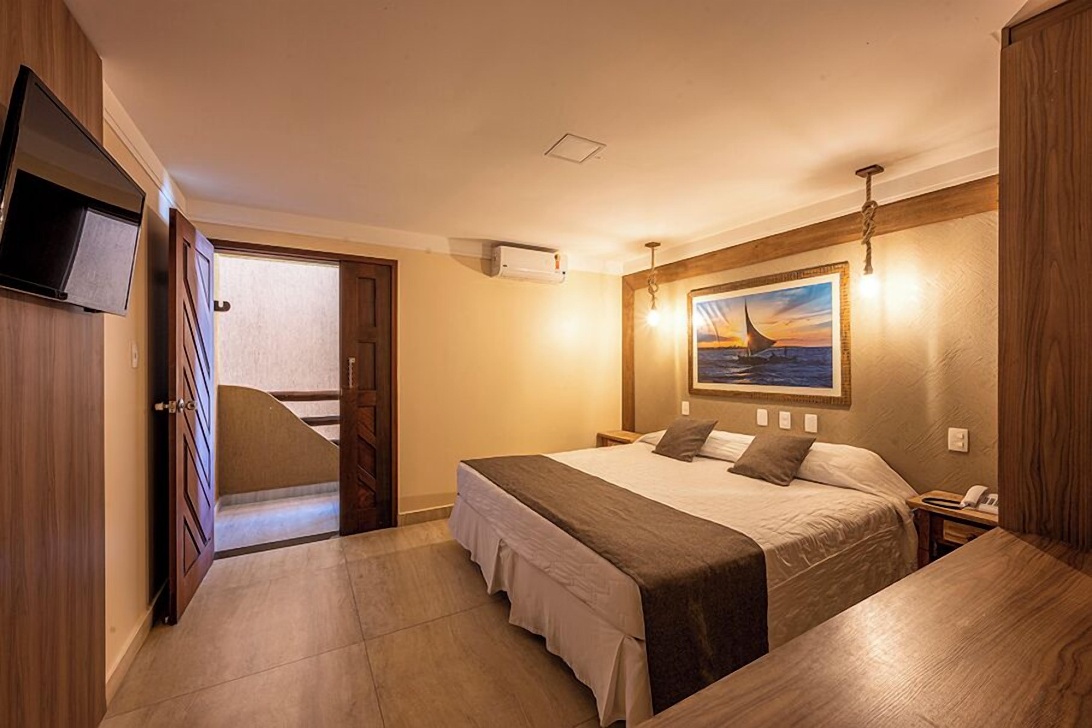 Quarto de hotel com cama, mesa de cabeceira e televisão fixada na parede.