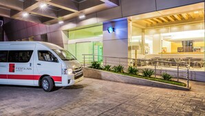 Una furgoneta blanca amb la marca 'Fiesta Inn' estacionada davant d'un edifici amb façana de vidre i zona d'exterior amb seients.