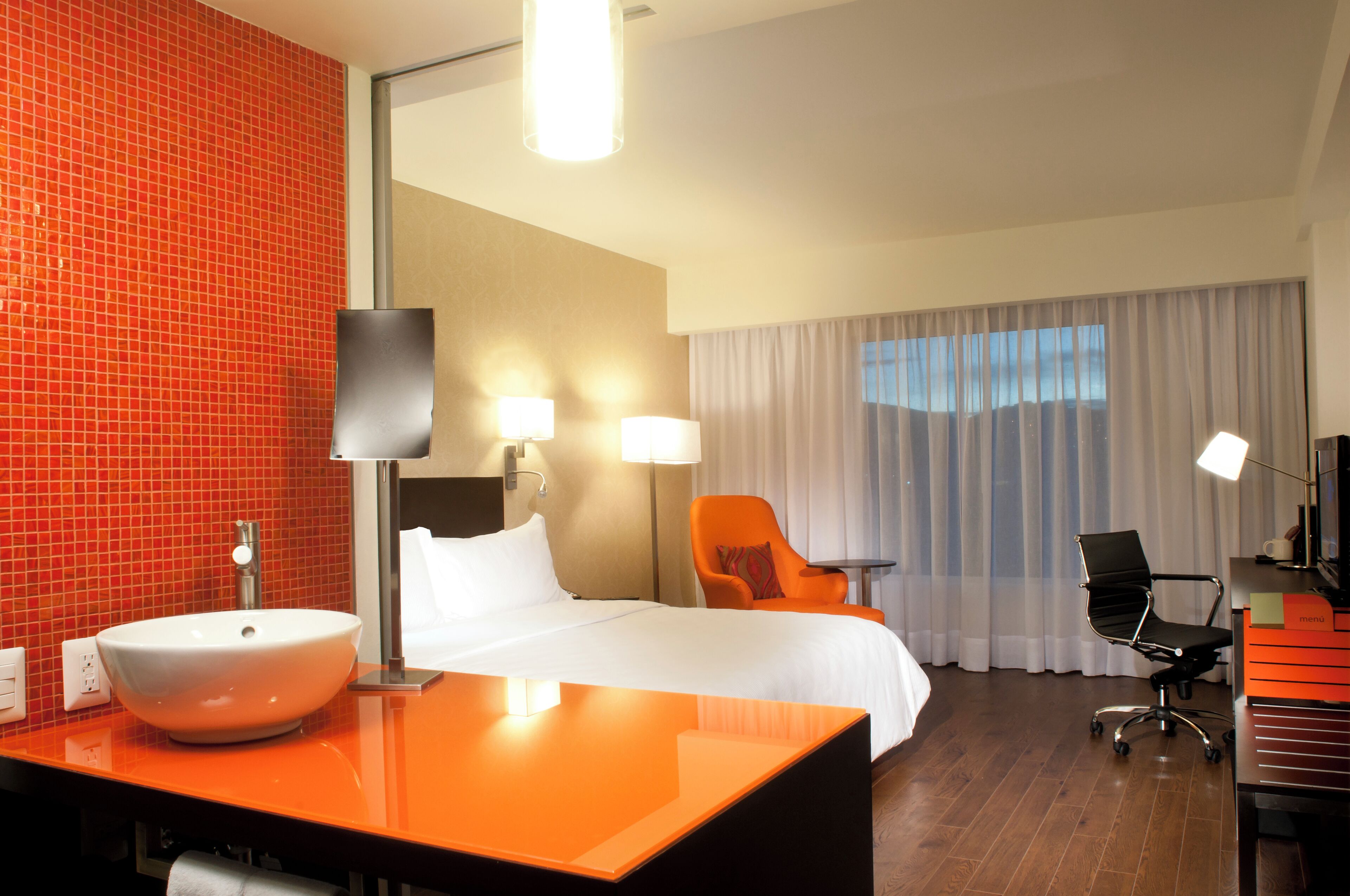 Habitación de hotel con una cama grande, un escritorio con una computadora portátil, una silla, una lámpara de escritorio y un baño con lavabo y espejo.