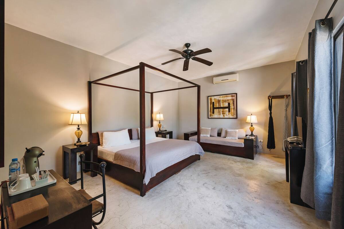 A four-poster bed in a hotel room with a ceiling fan, two bedside tables with lamps, a small table with a water bottle, and a mirror on the wall.