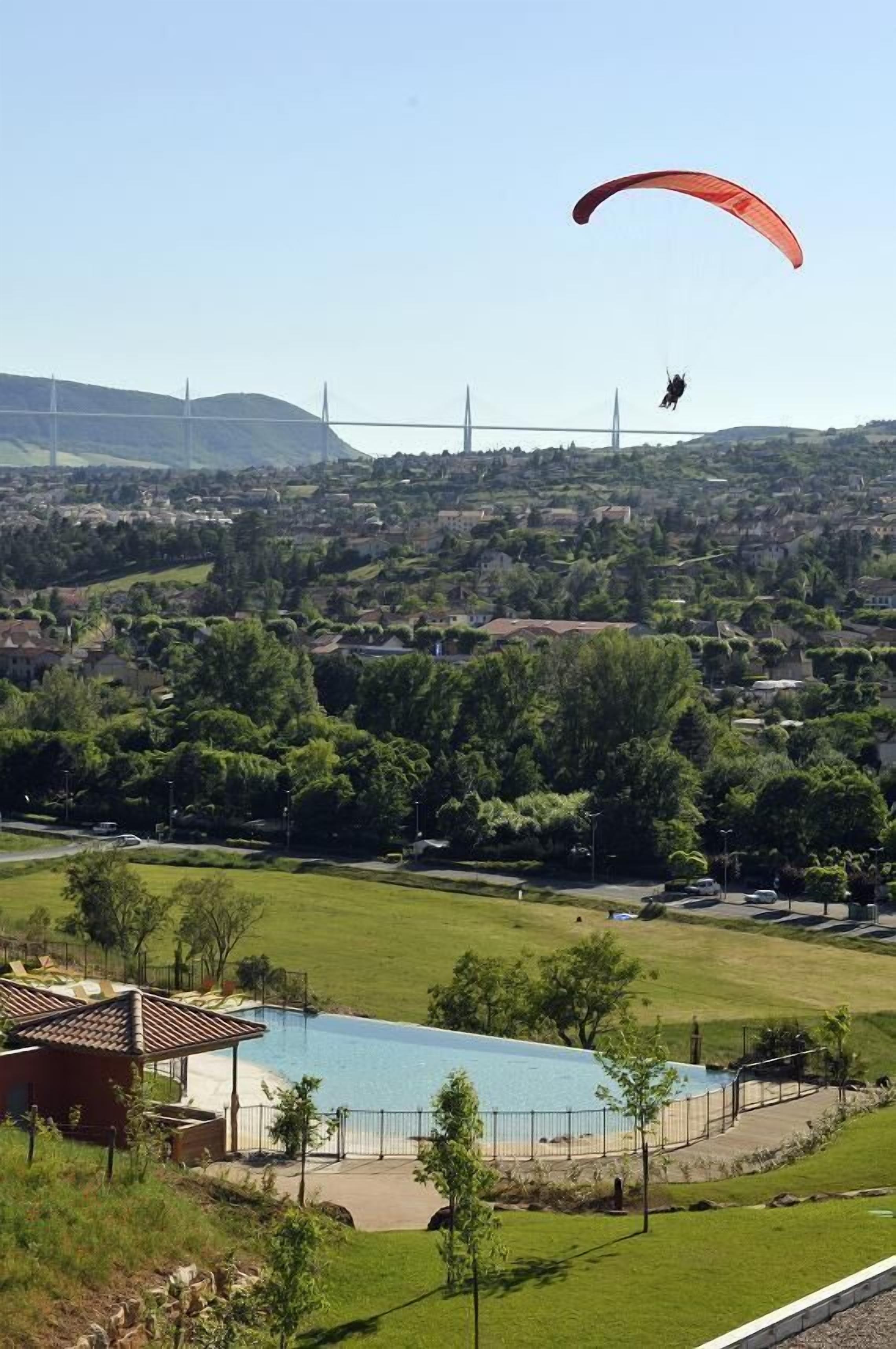 A person paragliding over a residential area with a large swimming pool and a bridge in the background.