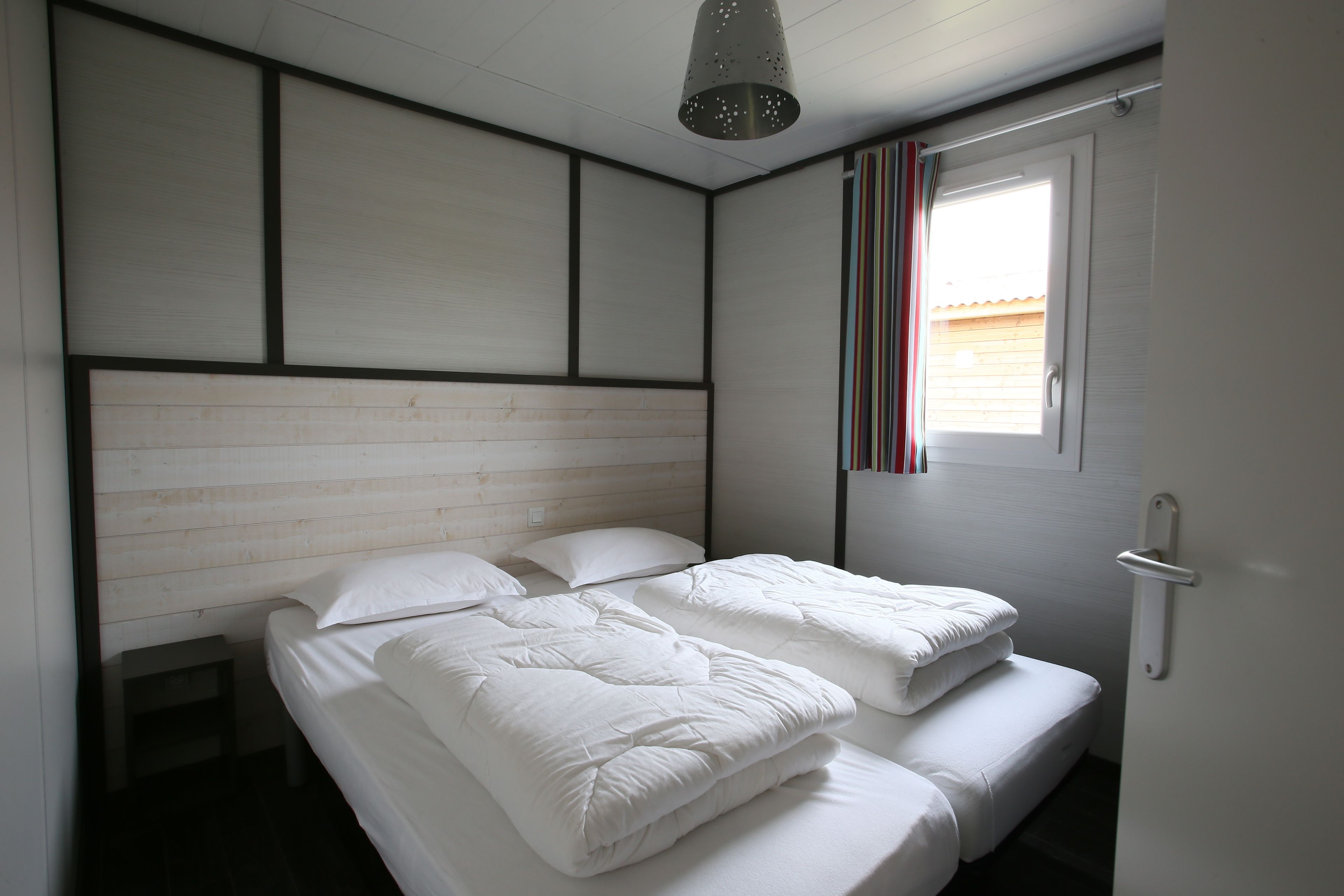 A bedroom with two beds, a wooden headboard, a window with striped curtains, and a ceiling light fixture.