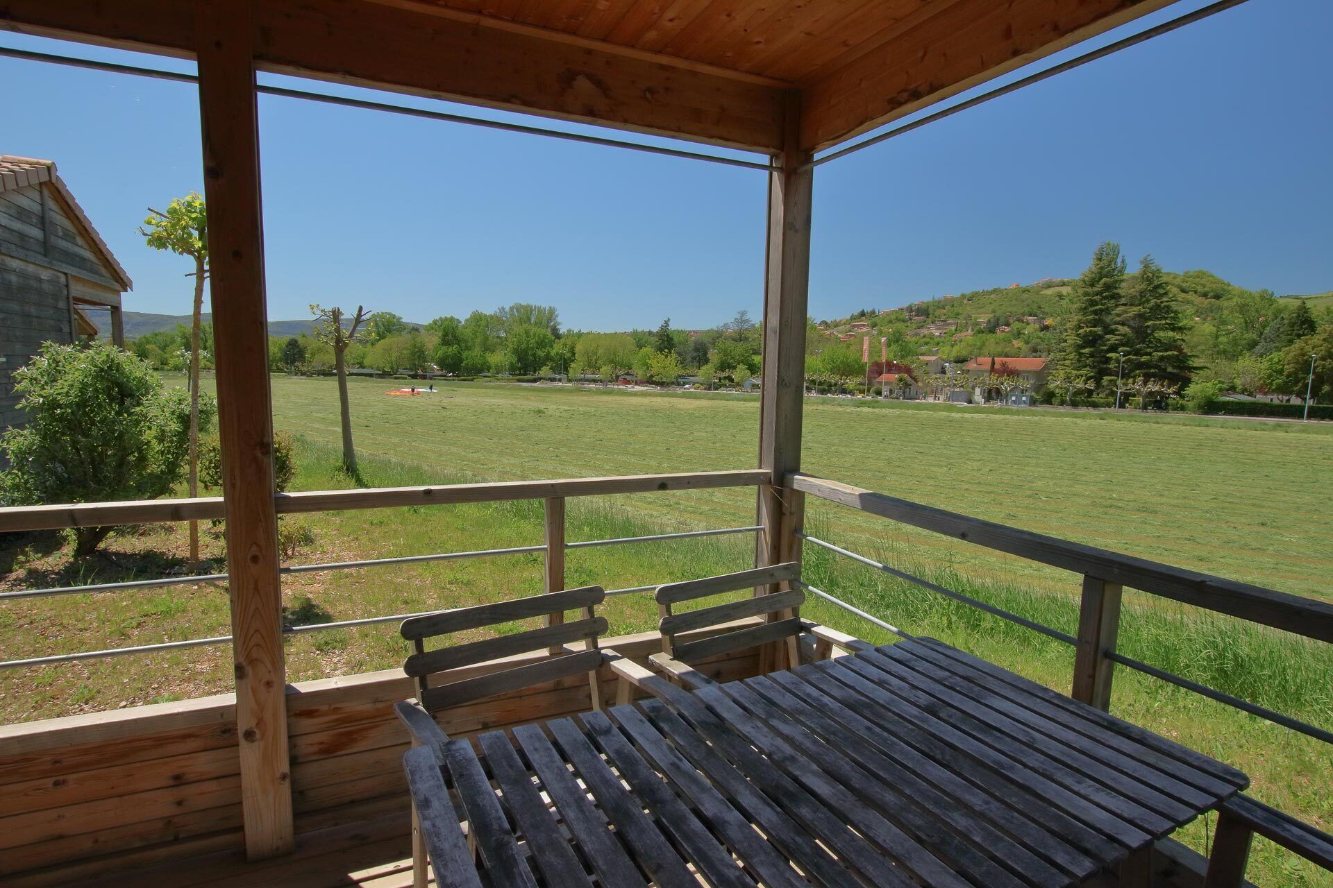 A covered outdoor seating area with a table and benches, overlooking a grassy field and distant buildings.
