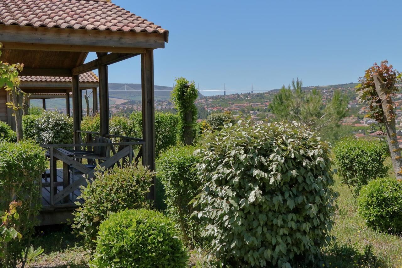 A wooden gazebo with a tiled roof overlooking a lush garden and a distant bridge.