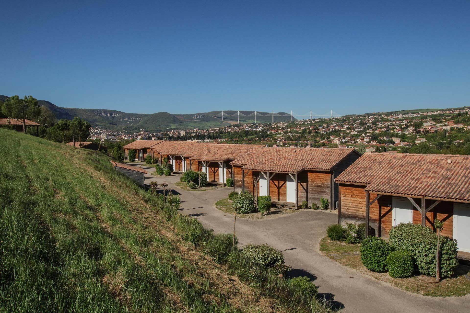 Une rangée de cabanes en bois aux toits de tuiles, construites sur une colline, d’où l’on peut admirer une ville et un pont situés au loin.