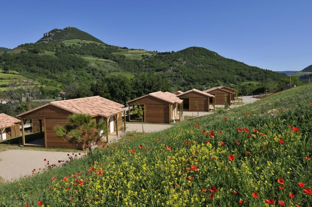 A row of wooden cabins with pitched roofs, set against a backdrop of green hills and a clear sky.
