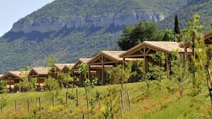 A row of wooden cabins with pitched roofs, set against a backdrop of green hills and a clear sky.