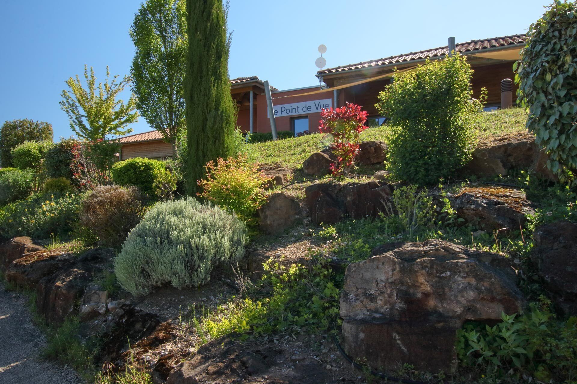 A building with a sign reading "le Point de Vue" surrounded by a landscaped garden with various shrubs and a prominent rock formation.