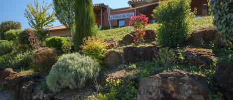 A building with a sign reading "le Point de Vue" surrounded by a landscaped garden with various shrubs and a prominent rock formation.