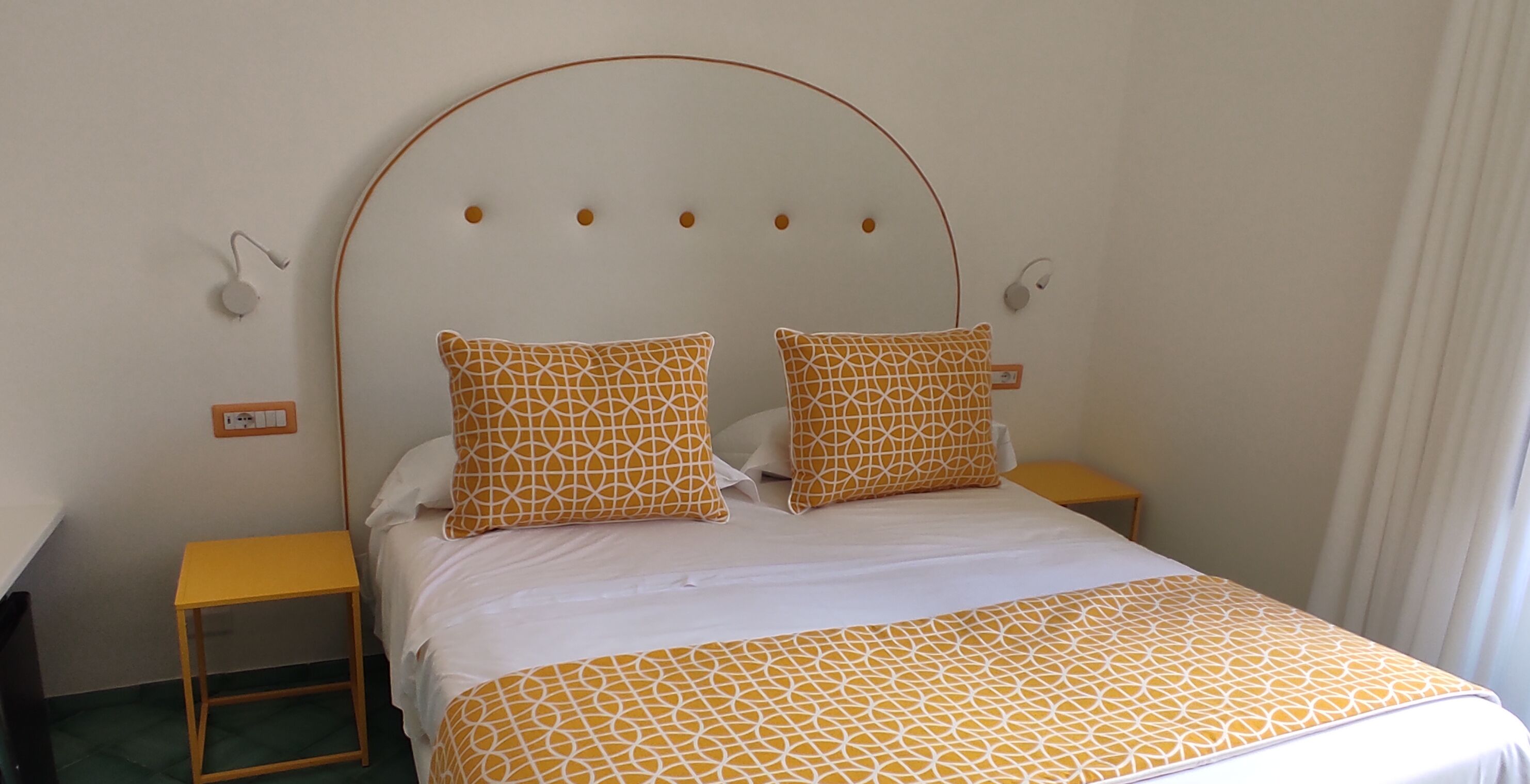 A neatly made bed with a geometric patterned bedspread and pillows, a wooden nightstand with a lamp, and a small yellow table beside the bed.