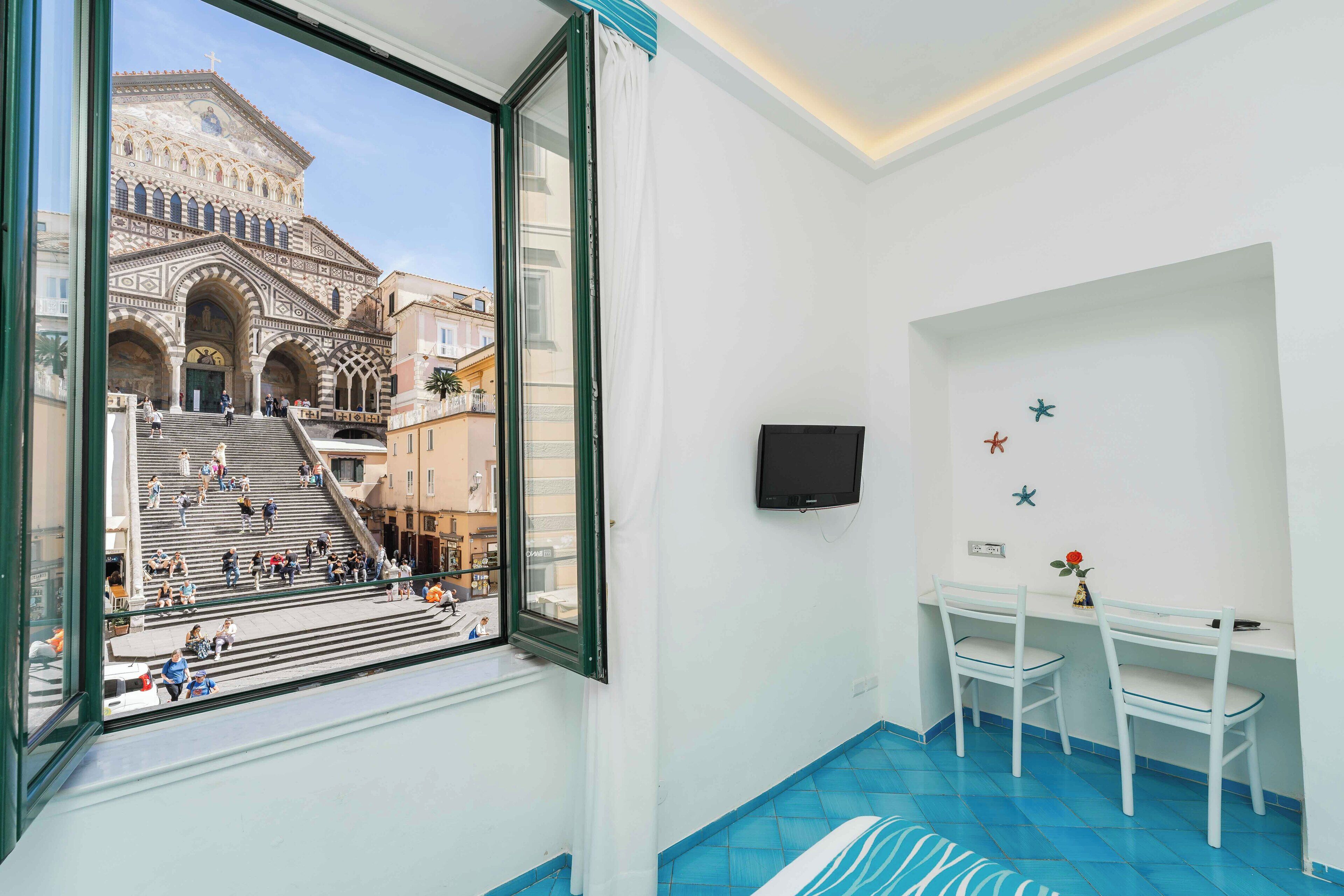 Una stanza con un'ampia finestra che si affaccia su un edificio storico con archi e gradini. È presente un tavolino con due sedie e un televisore fissato alla parete.