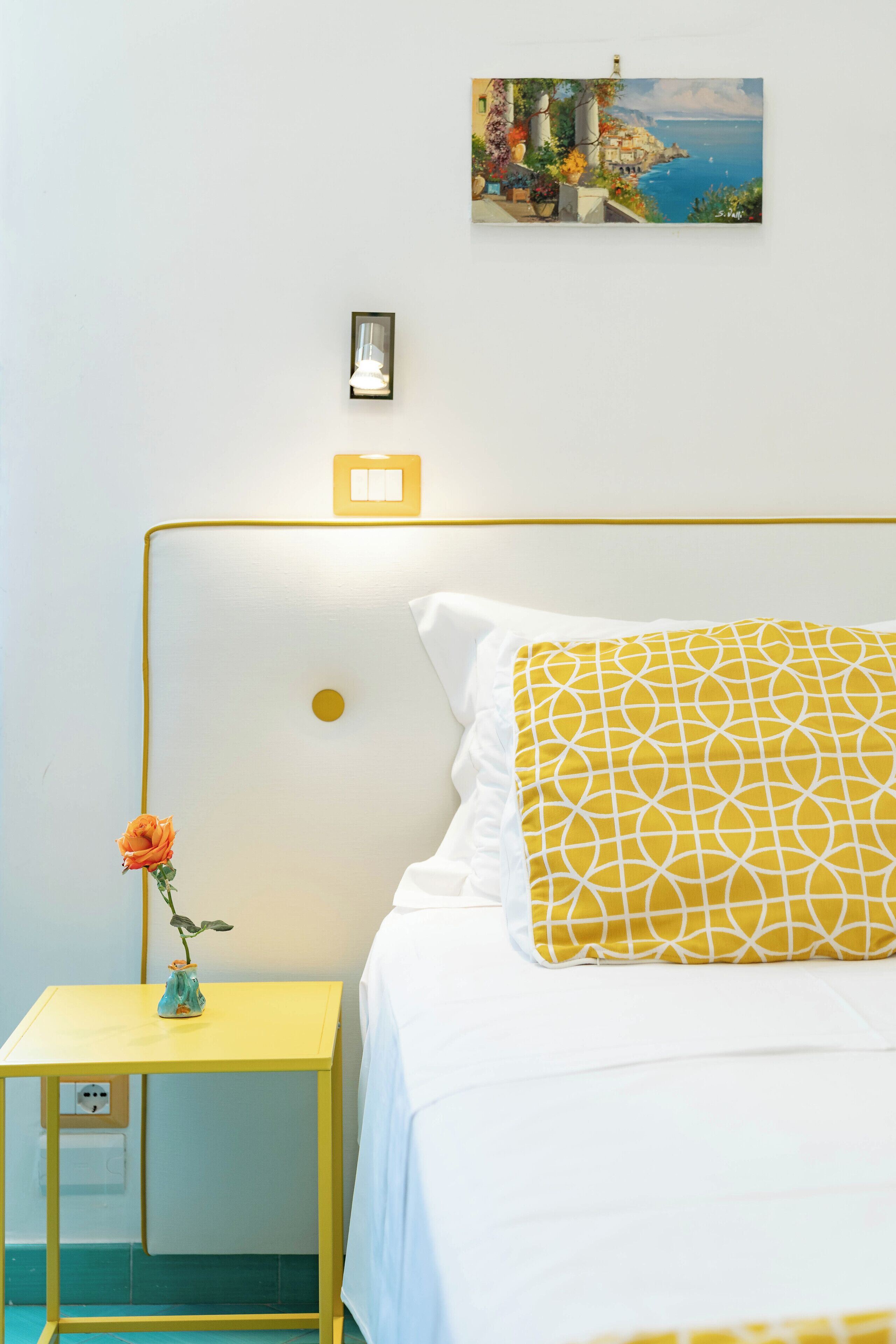 A neatly arranged bedroom with a bed, a bedside table with a vase, and a wall-mounted light.