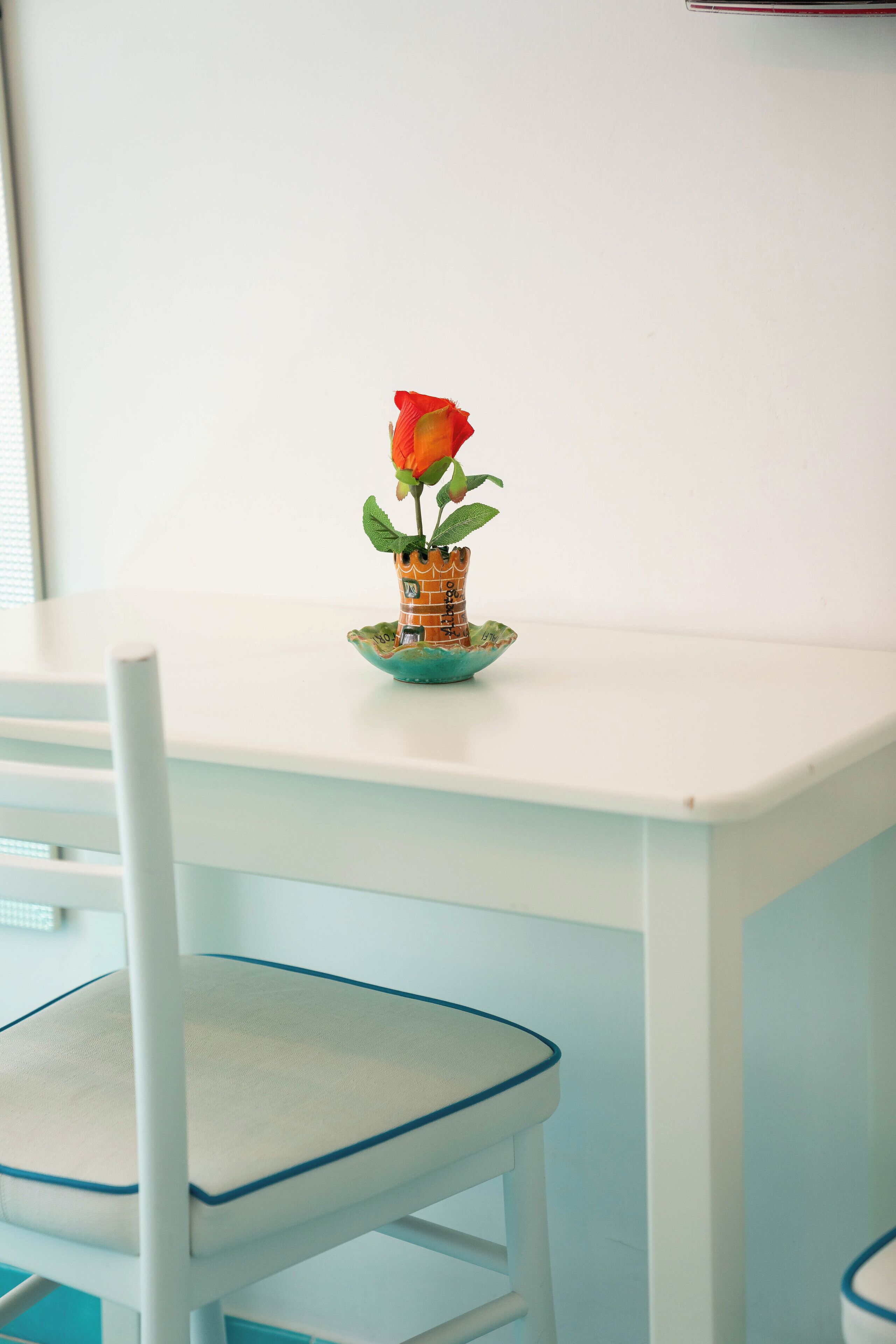 A white table with a small potted rose and a chair with a blue trim.