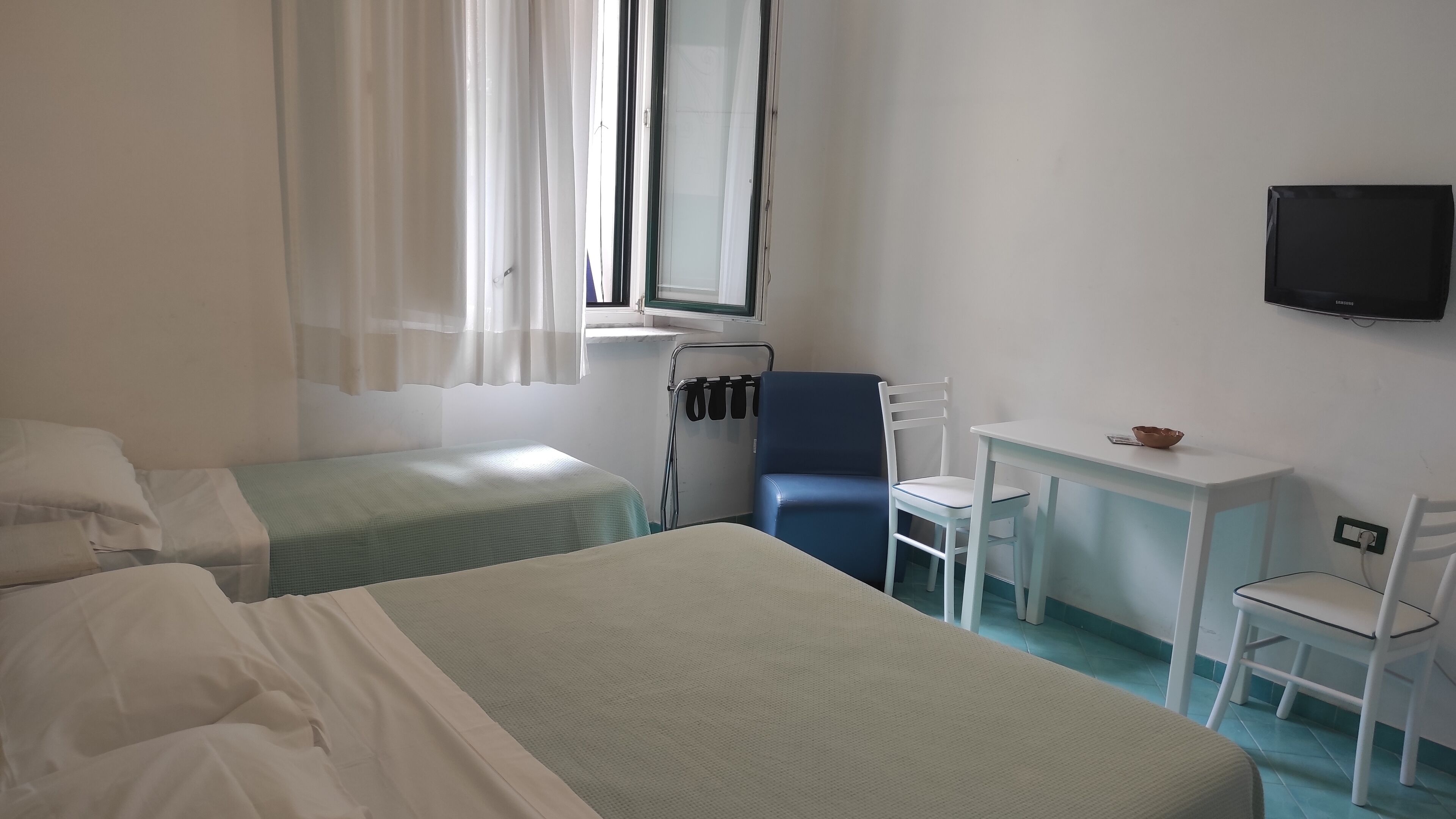 Une chambre d’hôtel avec deux lits simples, un petit bureau avec une chaise, un téléviseur fixé au mur et une fenêtre avec des rideaux.