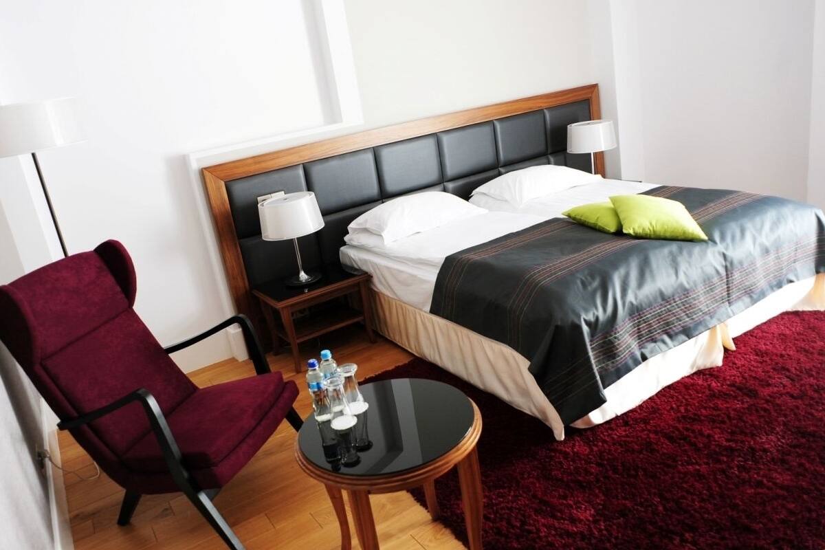 Una habitación de hotel con una cama, una silla, una mesita con botellas y una alfombra roja.