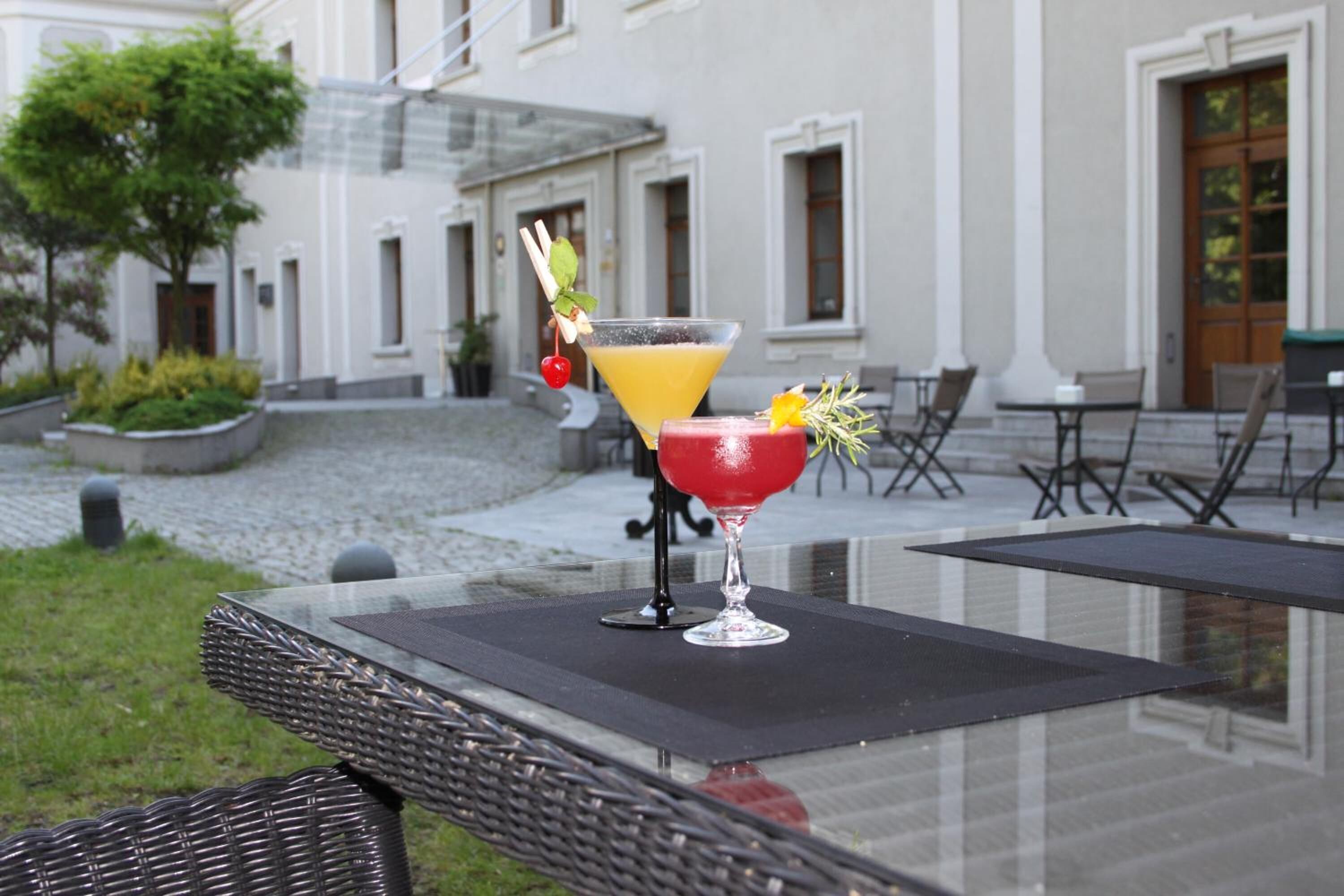 Pöydällä on kaksi juomaa, punainen ja keltainen cocktail, ja pöydän edessä on rakennus, jonka edessä on sisäpiha.
