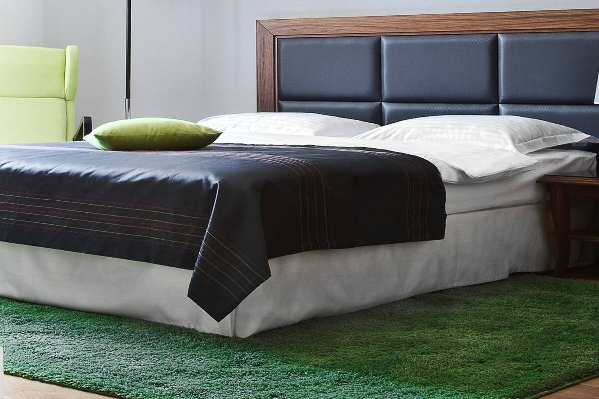 Una cama bien hecha con un cabecero oscuro, un almohadón verde y un almohadón blanco sobre una alfombra verde.