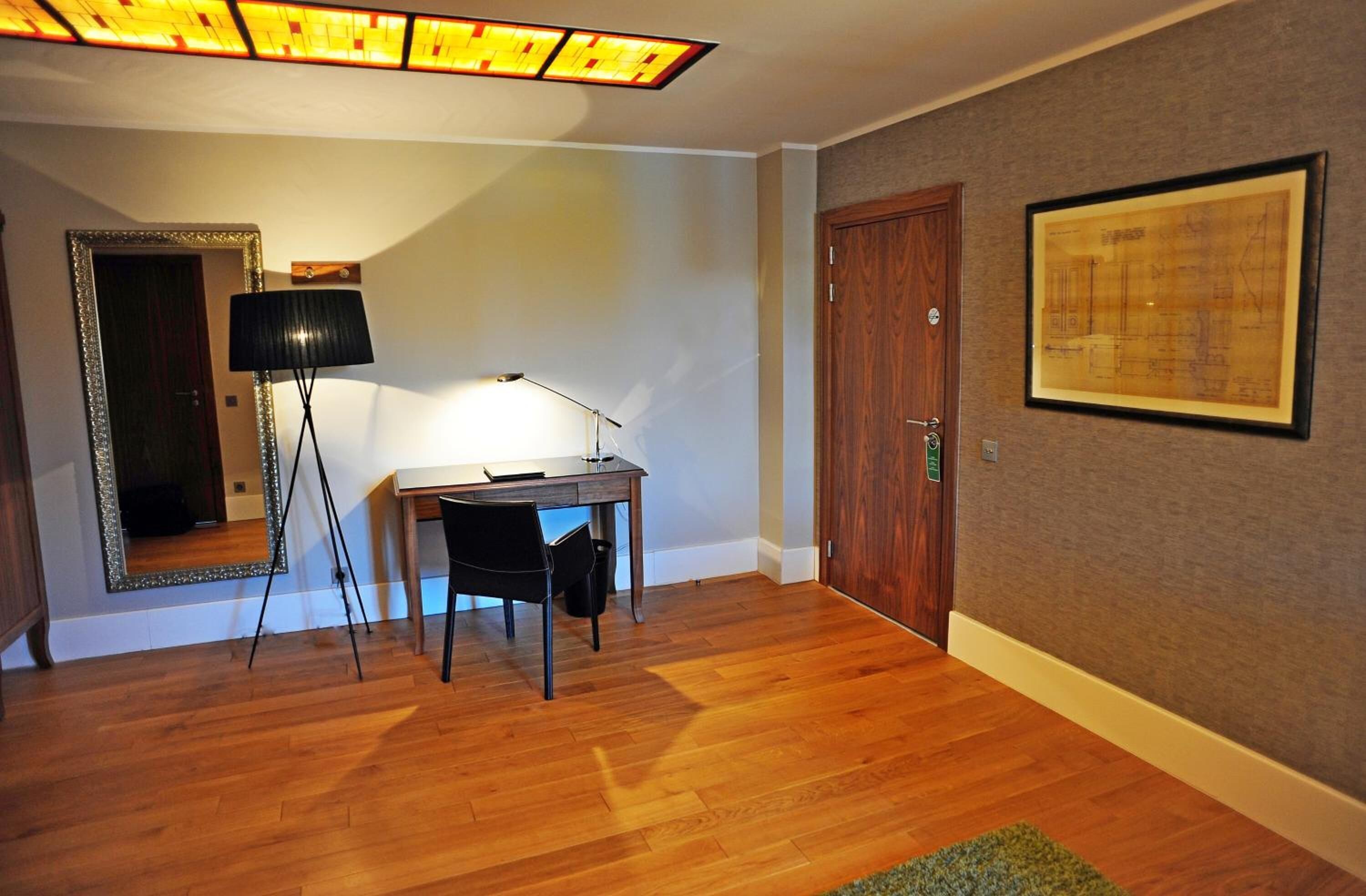 Una habitación con un escritorio de madera, una lámpara de pie, un plano arquitectónico enmarcado y una puerta.
