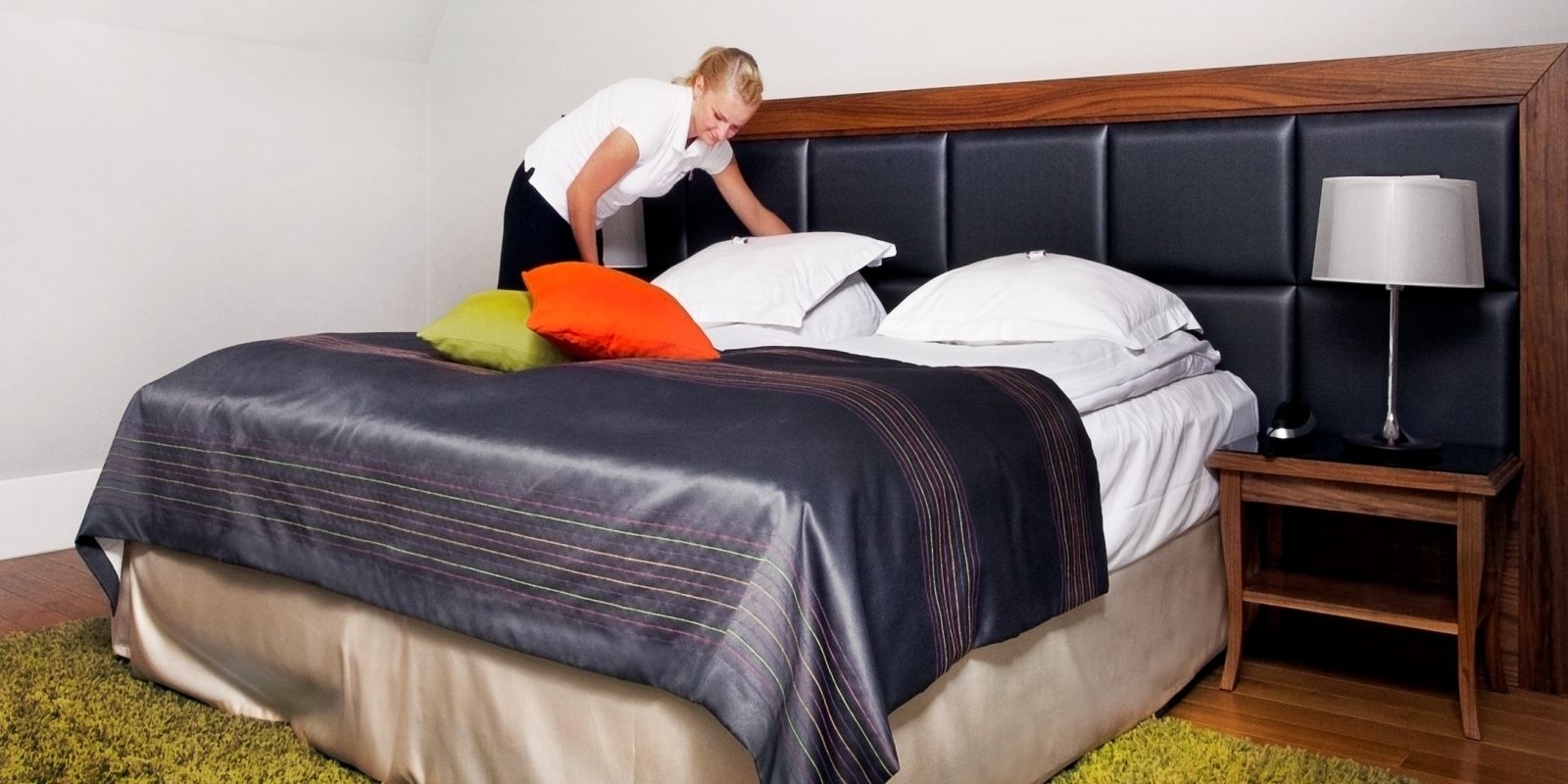 A neatly made bed with a dark headboard, a bedside lamp, and a green rug.