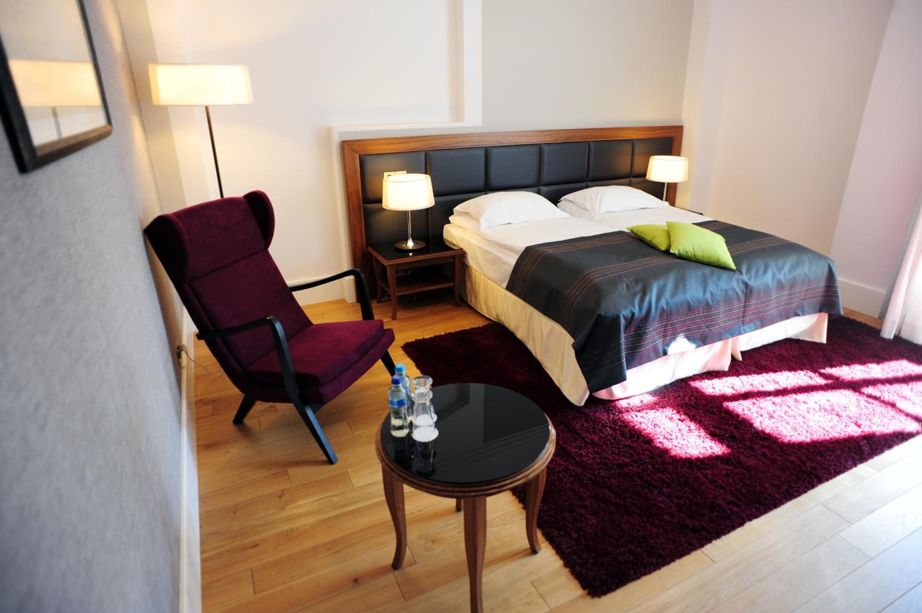 Habitación de hotel con cama, dos lámparas de noche, una silla, una mesita con botella y vaso, y una alfombra rojo oscuro.