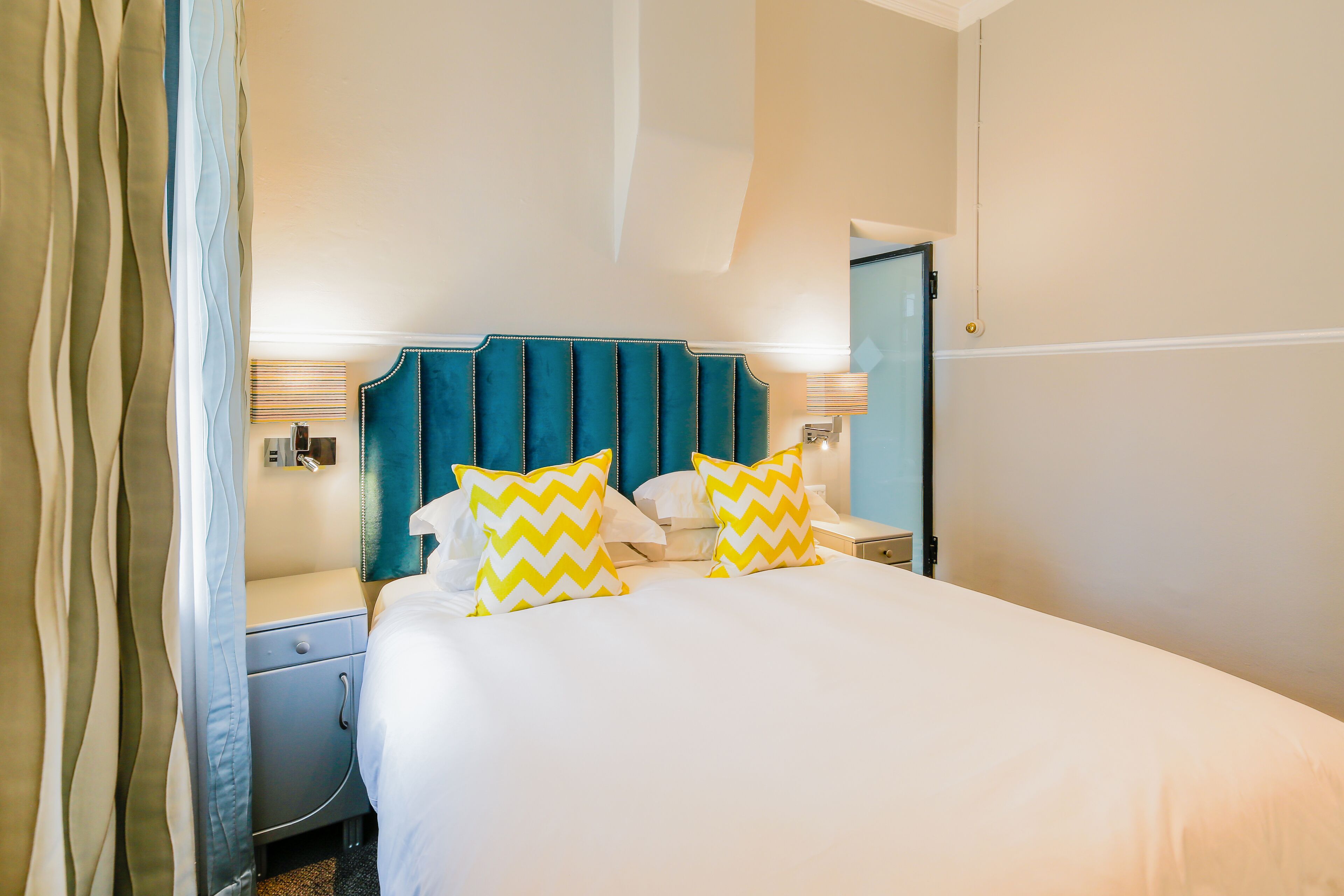 A hotel room with a bed, two nightstands, a teal headboard, and two yellow and white patterned pillows.