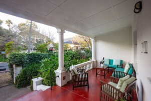 Un porche cubierto con piso rojo, columnas blancas y un área de descanso con muebles y cojines de madera.