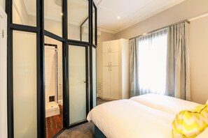A hotel room with a bed, a wardrobe, a bathroom with a shower, and a window with curtains. - Boschhuis Apartments - Stellenbosch (Stellenbosch Central)