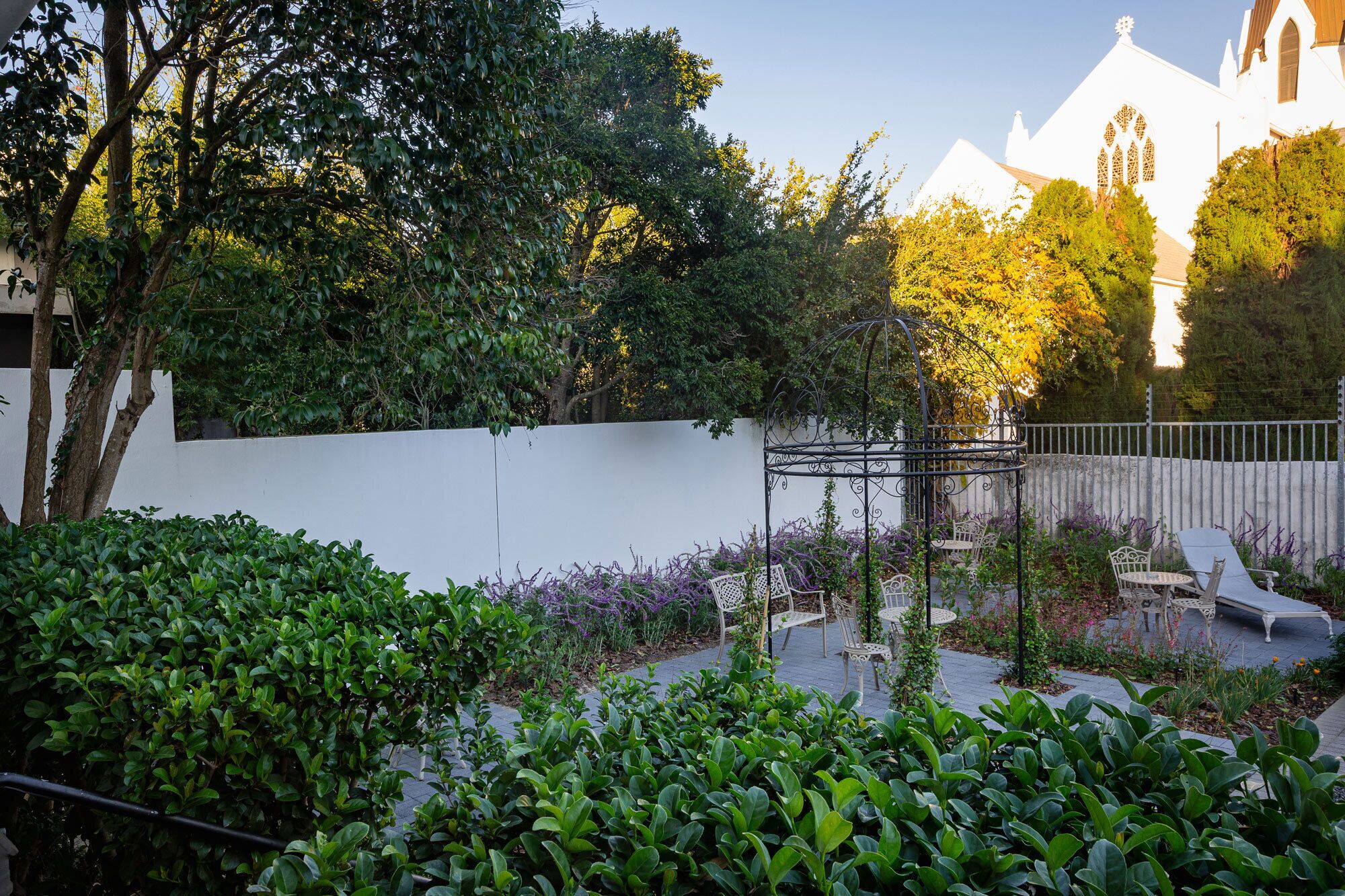 Un jardín con muro blanco, árboles y una pérgola de metal con sillas.