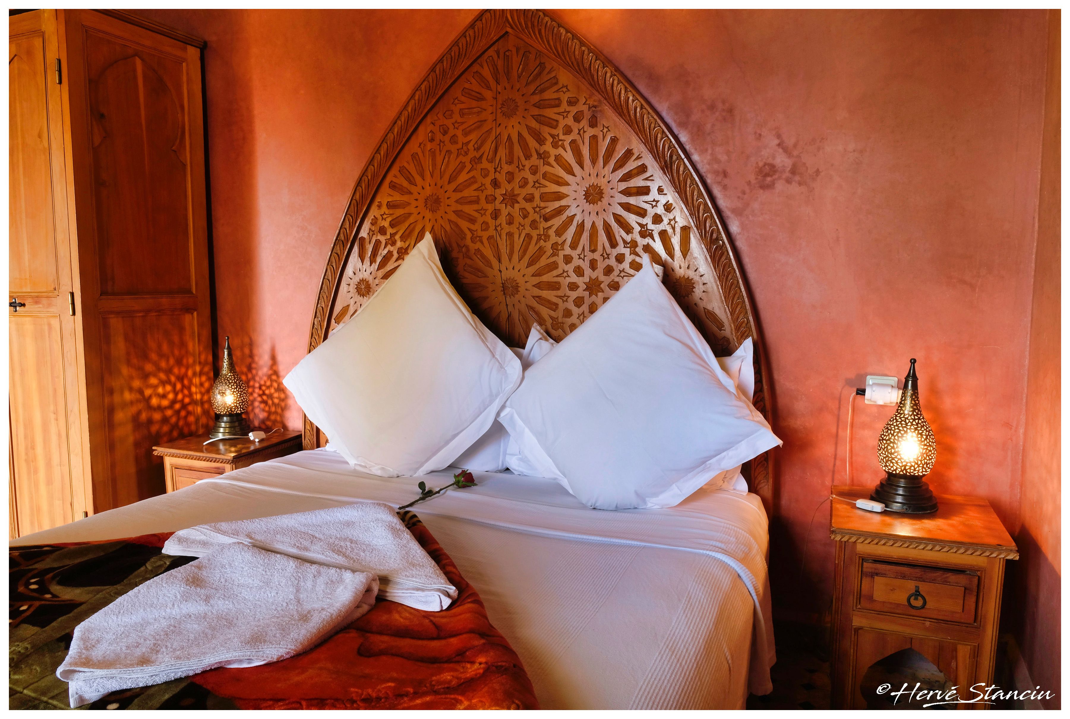 A neatly made bed with white linens and pillows, a red throw blanket, and a wooden headboard with intricate carvings. There are two bedside tables with lamps, one on each side of the bed.
