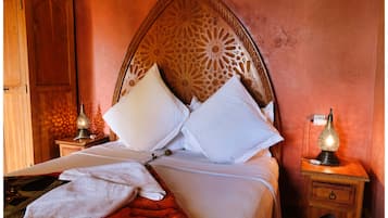 A neatly made bed with white linens and pillows, a red throw blanket, and a wooden headboard with intricate carvings. There are two bedside tables with lamps, one on each side of the bed.