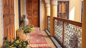 A corridor with ornate wooden doors, a large vase with yellow flowers, and a patterned red carpet.