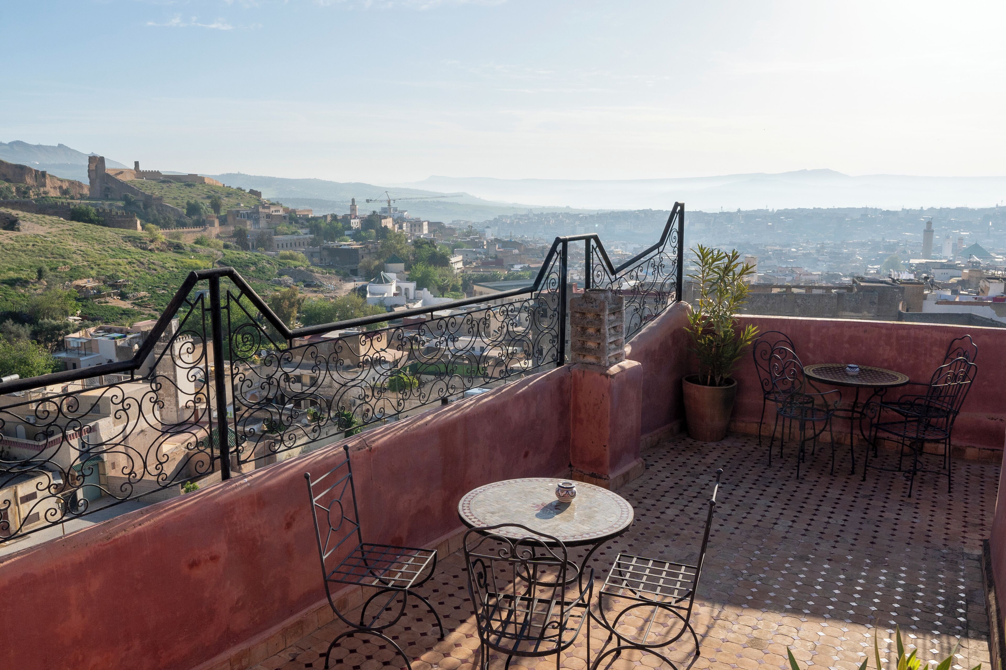 Una terraza en la azotea con una mesa redonda y sillas, una planta en maceta, y vistas a un paisaje urbano con edificios y colinas al fondo.