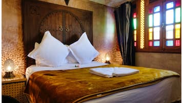 A bed with white pillows and a brown blanket, a wooden door with a circular design, and a window with colorful panes.