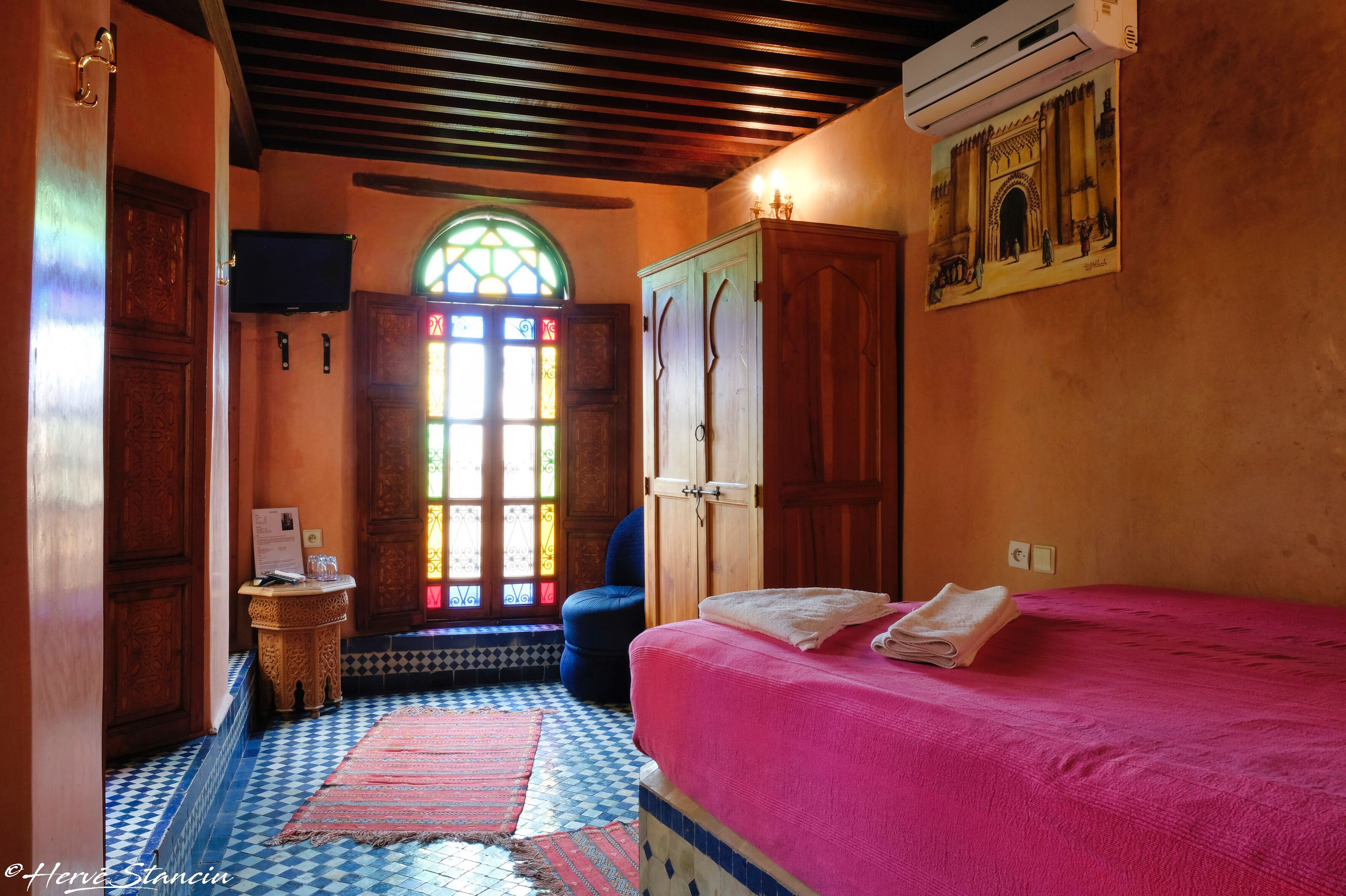 A room with a bed, a wooden wardrobe, a small table with a chair, a window with stained glass, and a tiled floor.
