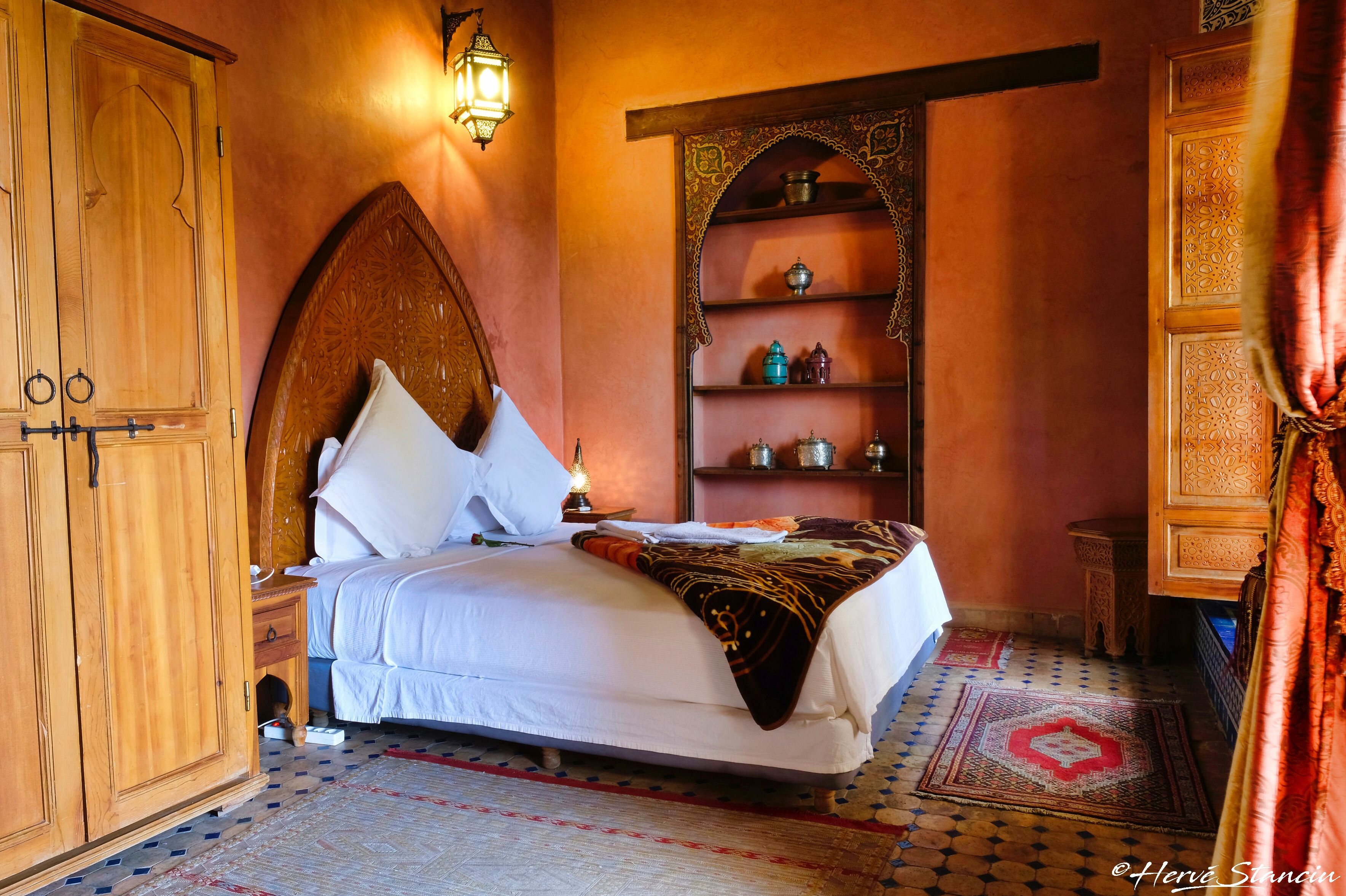 A traditional room with a bed, wooden wardrobe, shelves with decorative items, and a patterned floor.