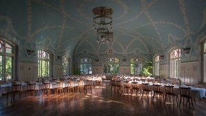 A large banquet hall with high ceilings and ornate ceiling patterns, large windows with stained glass, and rows of tables set for dining.